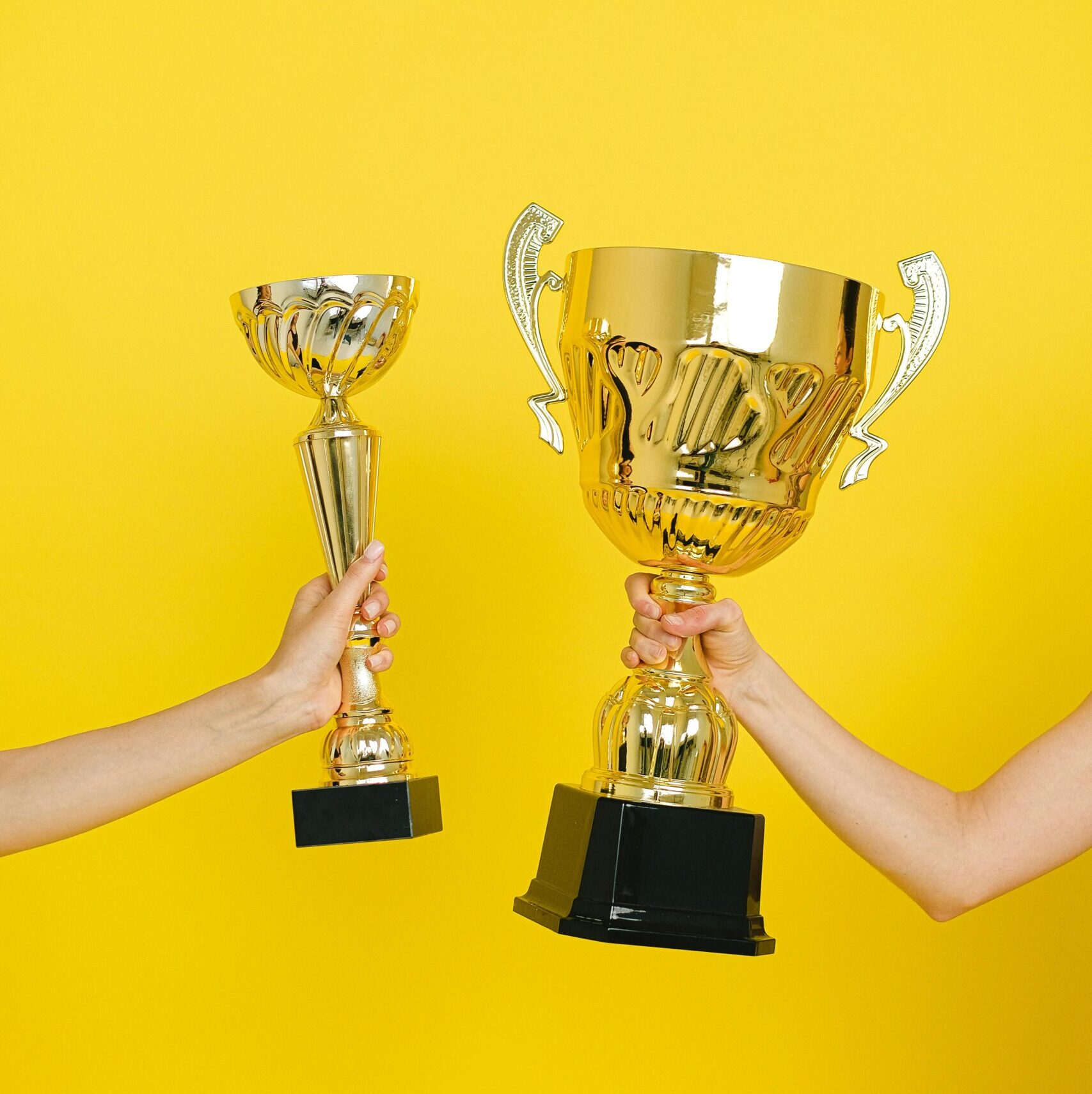 Two hands holding trophies against a vivid yellow backdrop, symbolizing success.